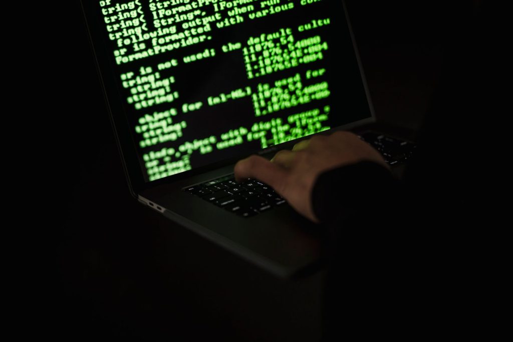 Hands typing on a laptop keyboard with green code on a dark screen